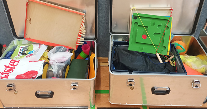 Zwei Koffer voller Bewegungs-Spielsachen für Kinder stehen in einer Sporthalle